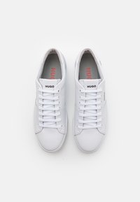 Białe skórzane sneakersy z okrągłym noskiem, płaskimi sznurowadłami i nazwą marki "HUGO" w czerni na języku, z teksturowaną gumową podeszwą.