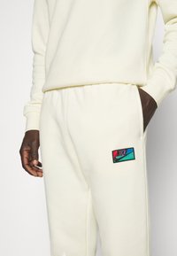 Cremefarvede Nike sweatpants med et broderet logo patch, der har røde, teal og sorte detaljer. Blødt stof med elastisk talje.