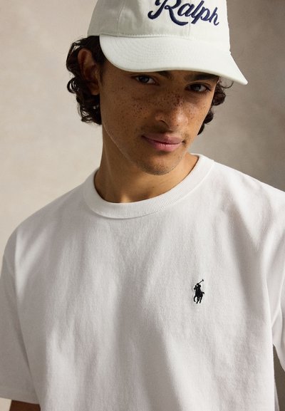 Jeune homme aux cheveux bouclés et taches de rousseur porte une casquette blanche Ralph et un t-shirt blanc avec petit logo polo noir, fond neutre.