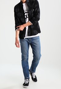 Camisa negra de botones con estampado de hojas sobre una camiseta blanca, combinada con jeans azul claro y zapatillas negras con cordones blancos.