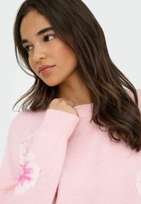 Pull léger rose fabriqué en tricot doux ; avec un motif floral sur les manches et un col et des poignets côtelés.