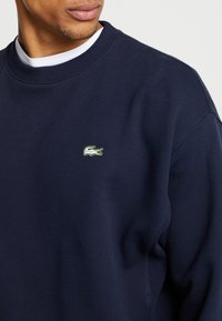 Sudadera de forro polar azul marino con cuello redondo y puños acanalados, con un pequeño logo de cocodrilo bordado en blanco y verde en el pecho.