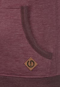Un sweat à capuche bordeaux avec une finition texturée, doté d'une poche avant et d'une étiquette en cuir carrée avec un logo. Le design comprend une bande en tissu contrastante.