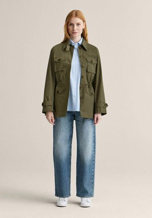 Kvinde iført en olivengrøn jakke med knapper over en lyseblå stribet skjorte, vide jeans og hvide sneakers, stående foran en ensfarvet baggrund.