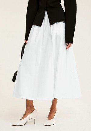 Kobieta w białej spódnicy midi, czarnym topie z długim rękawem, białych szpilkach i trzymająca czarną torebkę przy boku.