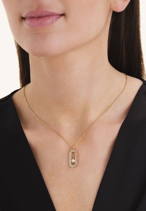 Donna che indossa una collana in oro con pendente ovale con piccola perla e pietre trasparenti, vestita con top nero.