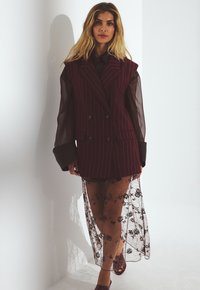 Gilet bordeaux a righe con design doppiopetto sopra una gonna in pizzo floreale nera trasparente. Include una blusa a maniche lunghe traslucida e calzature color marsala.