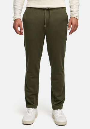 Mężczyzna w oliwkowych spodniach ze ściągaczem i białych trampkach, stojący na jednolitym jasnym tle.