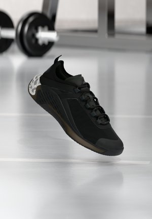 Chaussure de sport noire avec semelle sombre flottant au-dessus d'un sol de gym réfléchissant, avec une barre de musculation floue en arrière-plan.