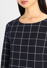 Haut bleu marine avec un motif à carreaux blancs. Présente un col rond et des manches longues, fabriqué dans un tissu lisse et texturé.