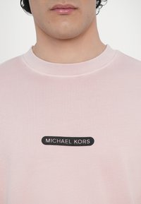 T-shirt de algodão rosa claro com decote redondo e um rótulo retangular preto com texto branco a dizer "MICHAEL KORS."