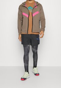 Bruine zip-up jack met roze accenten, bruin T-shirt, zwarte gebloemde shorts, zwarte leggings en lichtgroene sneakers met rode zolen.