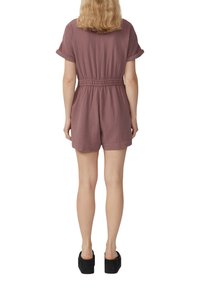 Kurze, mauvefarbene Jumpsuit mit gestepptem Taillenbereich, elastischem Bund und kurzen Beinen. Getragen mit schwarzen Plateauschuhen. Strukturierter Stoff.