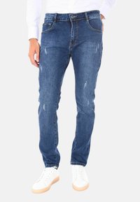 Jeans in denim blu con una vestibilità slim, leggeri segni di usura, design a cinque tasche e abbinati a sneakers bianche su uno sfondo neutro.