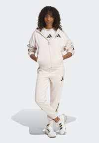 Lichtroze zip-up hoodie met zwarte accenten, bijpassende broek en witte sportschoenen met zwarte strepen. Krullend haar, staande pose.