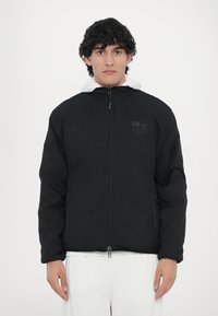 Black puffer jacket with a textured fabric, zip closure, and a white hood underneath. Features a small logo on the upper left side.
