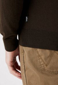 Maglione a maglia marrone con polsini e orlo a coste. Piccola etichetta nera sul lato. Lo sfondo presenta un tessuto beige chiaro dei pantaloni.