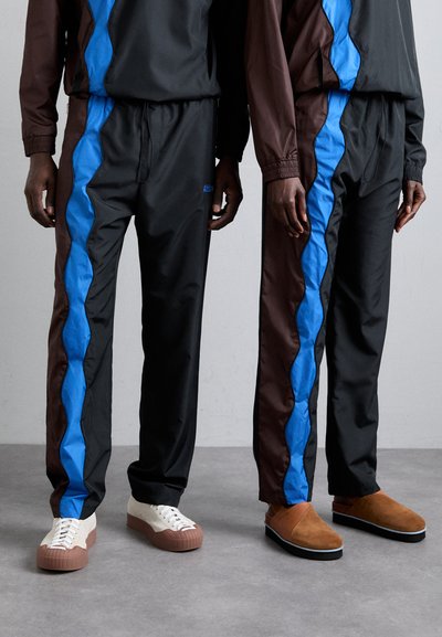 Chándales en negro y marrón, con franjas azules onduladas, fabricados en un tejido suave. El calzado incluye zapatos crema y marrones, así como zapatillas marrones.