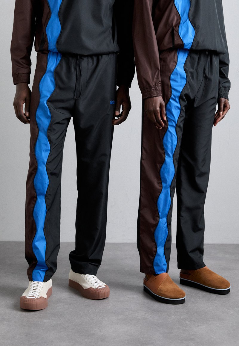 Des survêtements noirs et bruns, avec des rayures bleues ondulées, fabriqués en tissu lisse. Les chaussures comprennent des chaussures crème et brunes ainsi que des pantoufles brunes.
