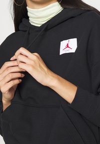 Zwarte hoodie met trekkoord, voorzien van een wit embleem met een rood logo. Gelaagd met een lichtgroene coltrui eronder. Soepele textuur.