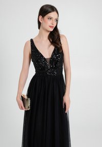 Schwarzes Abendkleid mit tiefem V-Ausschnitt, paillettenbesetztem Oberteil, Tüllrock und tailliertem Schnitt. Das Model hält eine kleine goldene Clutch.