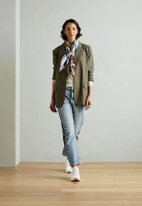 Blazer verde oliva su una maglietta beige, jeans azzurri e sneakers bianche. Una sciarpa a quadri multicolore è annodata attorno al collo.
