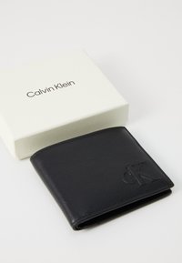 Portefeuille en cuir noir avec logo "ck" embossé à côté d'une boîte blanche de la marque Calvin Klein sur une surface blanche.