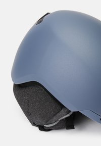 Smučarska čelada z matirano svetlo modro zunanjostjo, mehkimi črnimi ušesnimi blazinicami in elegantnim, zaobljenim dizajnom; ima prezračevalne reže na vrhu.