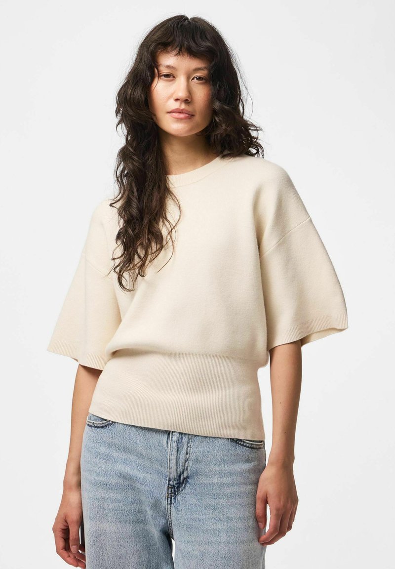 Femme aux longs cheveux foncés portant un pull crème à manches courtes et un jean bleu clair sur un fond clair uni.