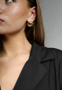 Pendientes de aro de oro con diseño texturizado y estriado. Forma circular, con intrincados detalles. Se llevan con una chaqueta negra.