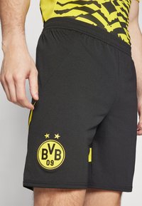 Man som bär svarta sportshorts med gult Borussia Dortmund BVB 09-logotyp och matchande gul och svart mönstrad skjorta.
