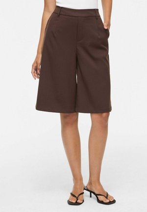 Femme portant un short ample marron foncé à la hauteur des genoux et des sandales à talons noires à brides, debout sur fond blanc.