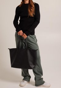 Svart tote-väska i strukturerad läder med två långa handtag. Bärs med lösa, gröna byxor och vita sneakers mot en neutral bakgrund.