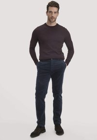 Dunkellila langärmliger Pullover, marineblaue Cordhosen und dunkle Schuhe. Das Model steht mit den Händen in den Taschen vor einem hellen Hintergrund.