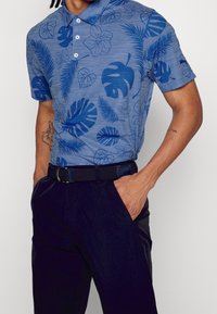 Sininen poolopaita tummansinisillä kukkakuosisilla, lyhyet hihat ja kaksinappinen kaulus; yhdistetty tummiin housuihin ja teksturoituun mustaan vyöhön.