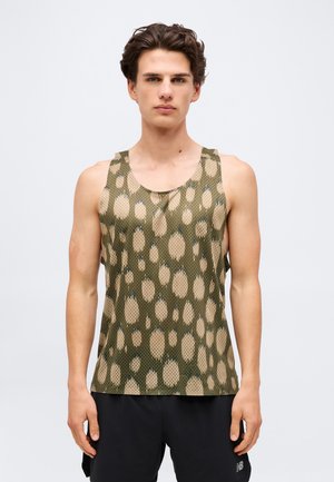 Junger Mann mit kurzen braunen Haaren, der ein ärmelloses olivgrünes Netz-Top mit beigen abstrakten Flecken und schwarze Shorts trägt.