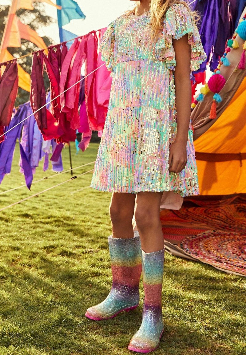 Enfant portant une robe pastel à sequins et des bottes de pluie arc-en-ciel scintillantes, debout sur l'herbe près de bannières colorées en tissu et d'une tente décorée.