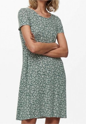 Mujer con los brazos cruzados, vestida con un vestido verde de manga corta con un patrón de pequeñas flores blancas, sobre un fondo liso.