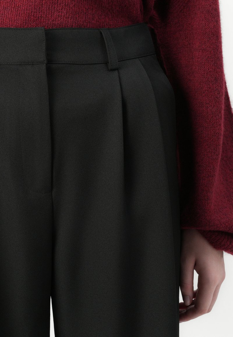 Hochgeschnittene schwarze Hose mit Falten, aus einem glatten Stoff gefertigt. Verfügt über einen Gürtel mit Schlaufen und sitzt oberhalb der Taille.