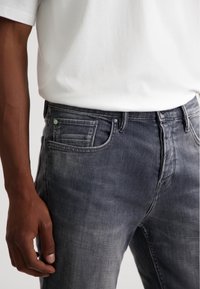 Jeans din denim gri cu un design clasic cu cinci buzunare, având o ușoară decolorare și un accent de buton albastru deschis pe buzunarul din față.