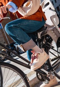 Béžové tenisky s růžovými šnuřovadly s vzorem, světle šedé ponožky, džínové kalhoty s manžetou a hnědá semišová bunda, sedící na kole.