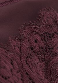 Primo piano di tessuto di pizzo color bordeaux con motivi floreali intricati e bordi smerlati su fondo liscio.