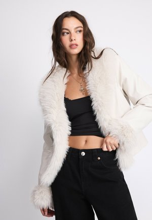 Jeune femme aux longs cheveux bruns portant une veste blanche bordée de fausse fourrure, un crop top noir et un pantalon noir sur un fond uni.