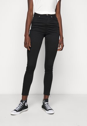 Persoon die zwarte high-waisted skinny jeans en zwarte hoge sneakers met witte veters draagt, staand tegen een eenvoudige witte achtergrond.