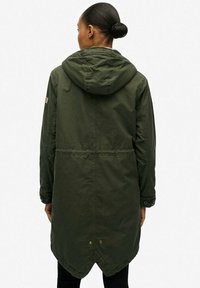 Parka de couleur vert olive avec une capuche, taille cintrée, manches longues et un ourlet arrière plus long. Présente un petit écusson avec un drapeau sur l'épaule gauche.