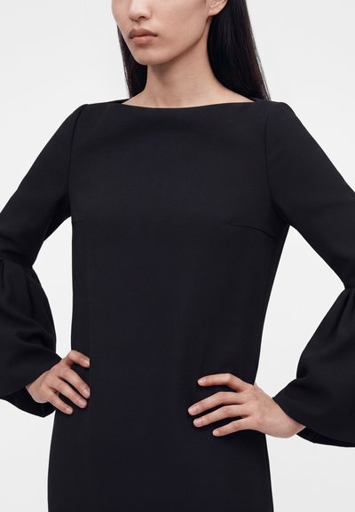 Γυναίκα με μαύρο φόρεμα με μακριά, φαρδιά μανίκια και τετράγωνη λαιμόκοψη, όρθια με τα χέρια στηρίγματα στους γοφούς μπροστά σε απλό φόντο.