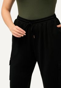 Černé joggingové kalhoty s elastickým pasem a nastavovací šňůrkou. Obsahují boční kapsy a mají měkkou, hladkou texturu.