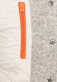 Grijze en witte jack van gevoerd materiaal, met een oranje rits en een zilveren drukknop. Geverfde grijze zijpaneel voor contrast.
