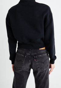 Felpa nera con colletto alto e polsini a coste, abbinata a jeans grigi a vita alta con etichetta in pelle e doppie tasche posteriori.