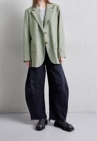 Blazer oversize verde chiaro con bottoni dorati, due tasche anteriori, abbinato a jeans larghi scuri con orli sfrangiati e stivali neri.
